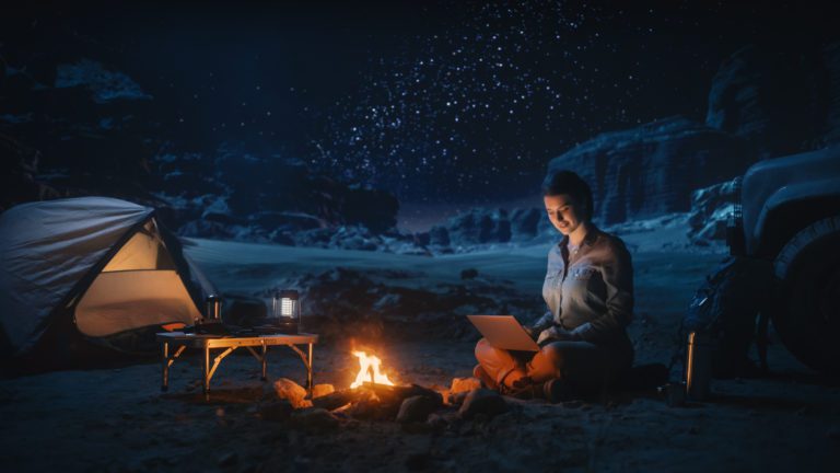 Night Tent Camping in Canyon: Female Traveler Uses Laptop Computer Sitting by Campfire. Woman on Digital Remote Work, e-shopping, ecommerce, Using Internet, Social Media Posting on Vacation Trip