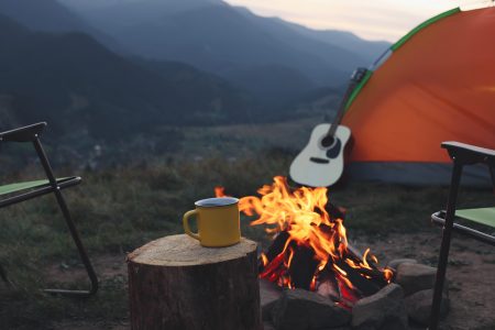 Yellow mug with hot drink on wooden stump near bonfire outdoors. Camping season